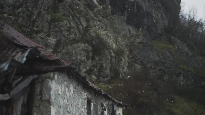 Light snow falls on a derelict stone building with a rusty tin roof at the base of a rocky cliff