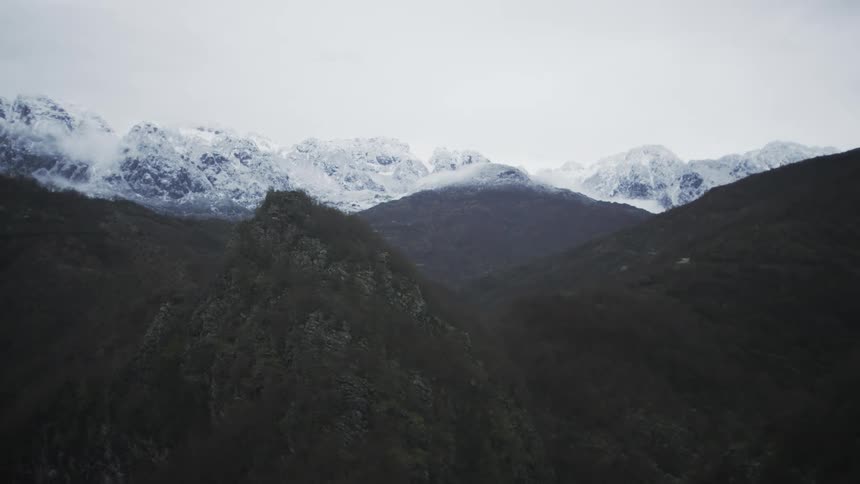 A slow aerial pan across a forested valley reveals a majestic snow-capped mountain range on a cloudy day