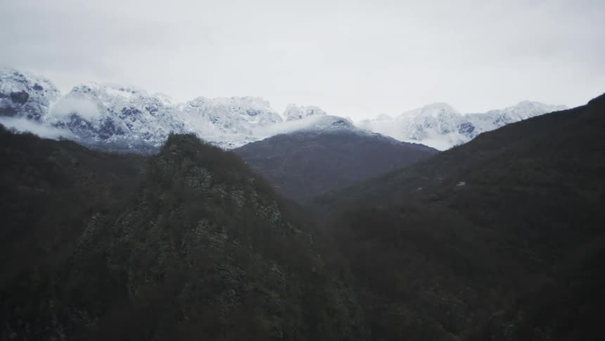 A slow pan across a vast, gloomy mountain valley reveals snow-capped peaks and a lone car driving along a remote, winding road