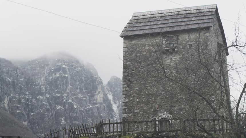 An ancient stone watchtower stands stoically against a backdrop of dramatic, snow-dusted mountains shrouded in mist