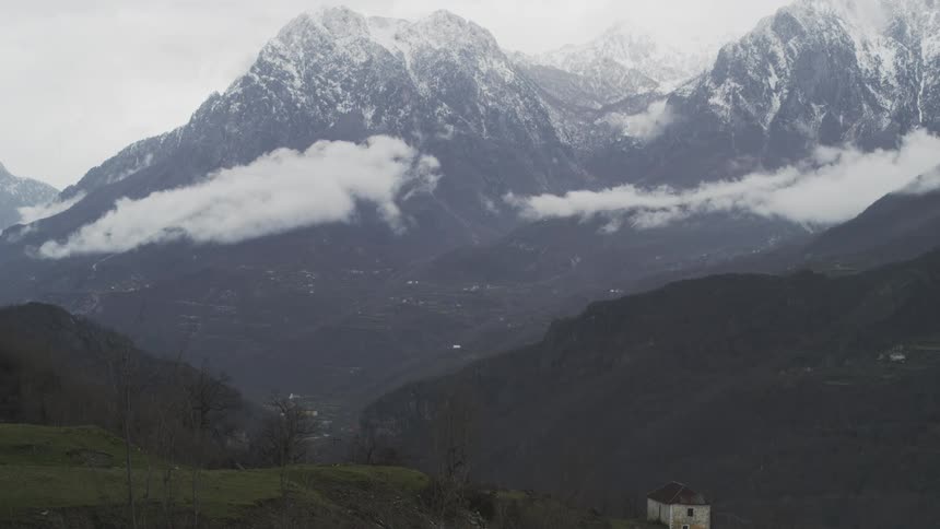 A static wide shot captures an isolated stone house in a valley below majestic snow-capped mountains shrouded in clouds on an overcast day