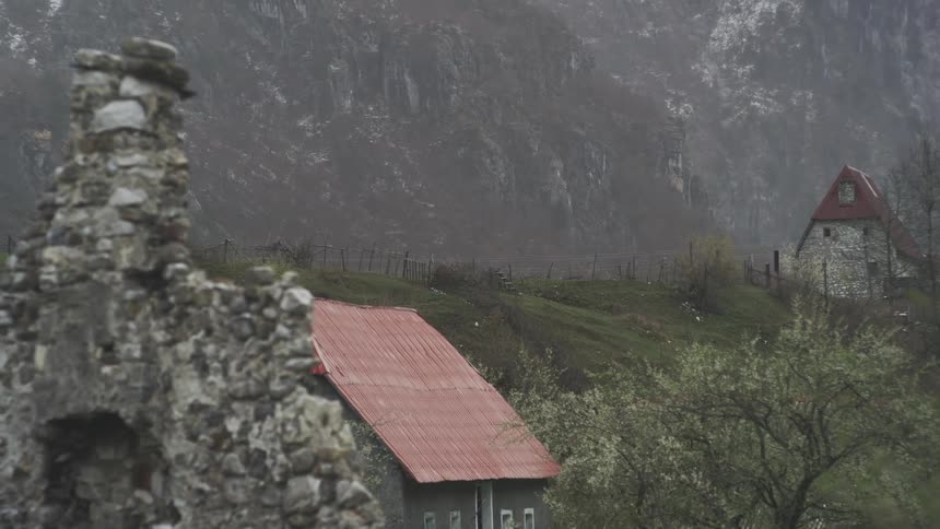 Rain falls gently on an old village with a red-roofed house and ancient stone ruins nestled in a misty mountain valley