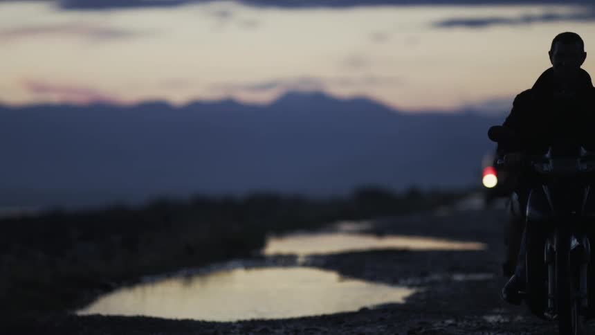 In slow motion, silhouetted bikers ride toward the camera on a desolate road at dusk, with distant mountains framing the scene