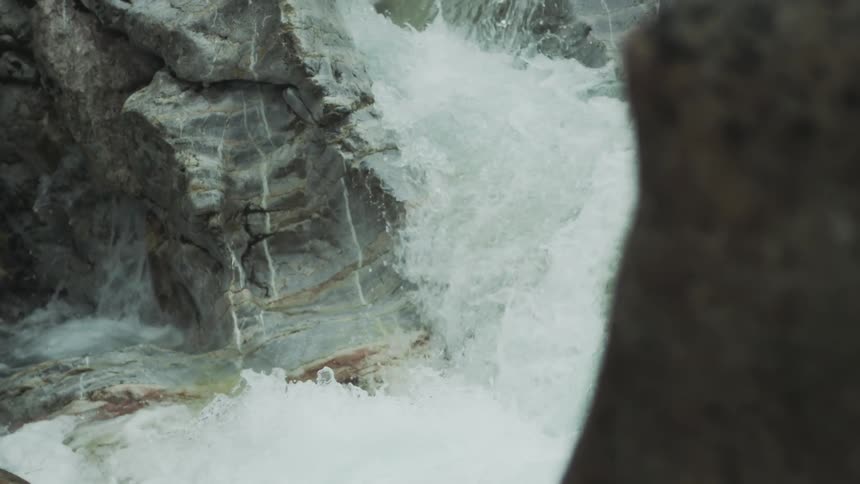 A powerful mountain stream crashes against ancient, layered rocks in a close-up slow-motion detail shot