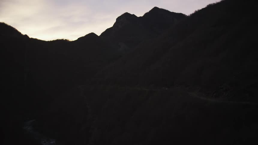 A lone car with its headlights on navigates a winding road carved into a vast, dark mountain range at dusk