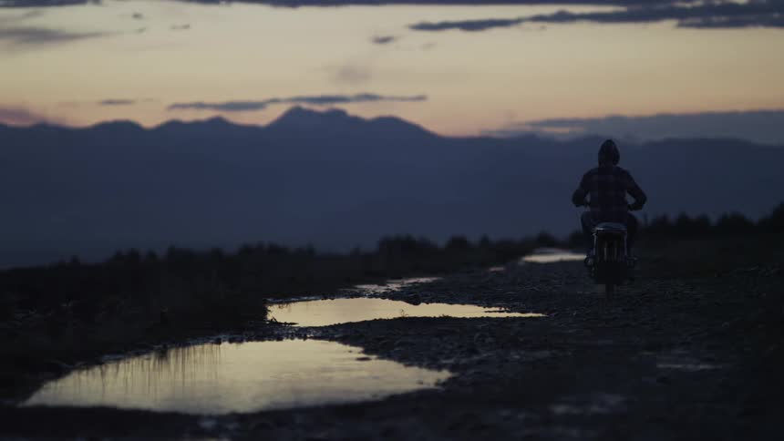 A lone motorcyclist rides on a dirt road towards a mountain range during a moody twilight