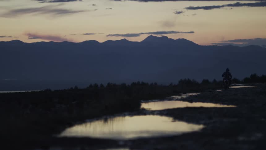 A lone motorcyclist sits on a muddy road at dusk, with puddles reflecting the beautiful mountain sunset
