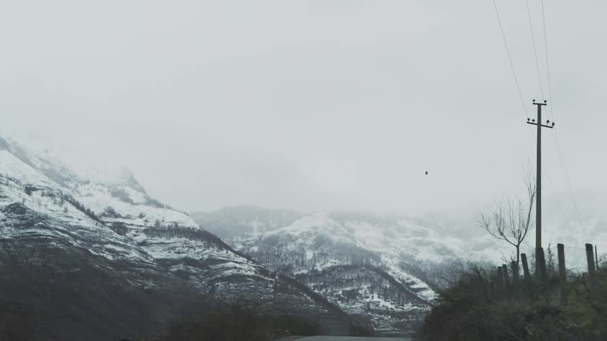 A point-of-view shot slowly advances along a rural road in a mountain valley, with snow-covered peaks emerging through the fog on a gloomy day