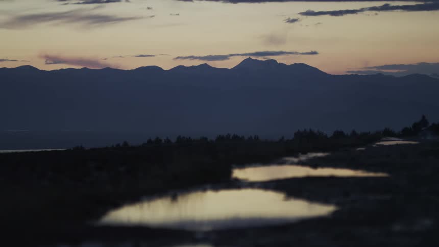 Puddles on a desolate trail reflect the soft twilight sky over a silhouetted mountain range at dusk