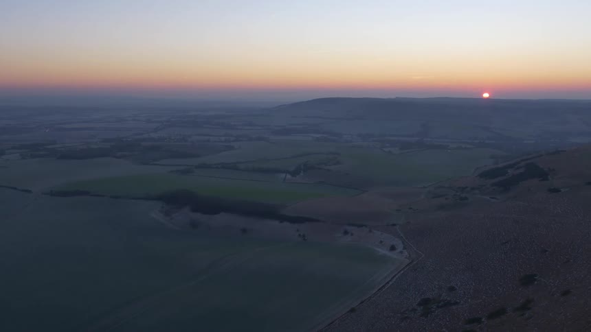 A serene aerial view shows the sun rising over a vast rural valley of rolling hills, with a frosty slope in the foreground