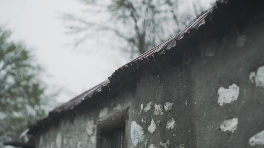Raindrops fall in slow motion on the rusty corrugated roof of an old stone shed on a gloomy day