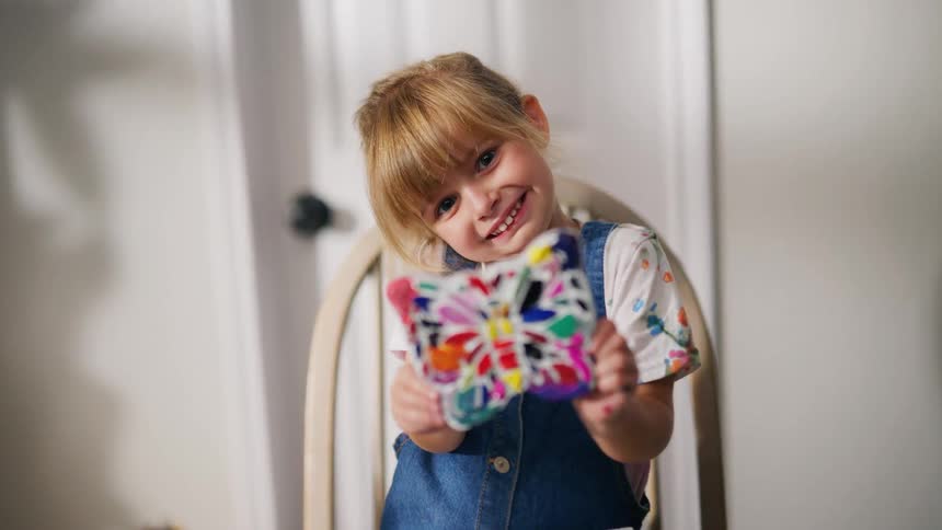 An adorable little girl with blonde hair proudly displays her colorful butterfly painting