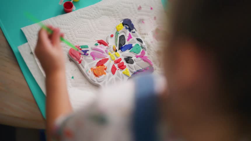 An over-the-shoulder view shows a little girl carefully painting a plaster butterfly craft with vibrant colors