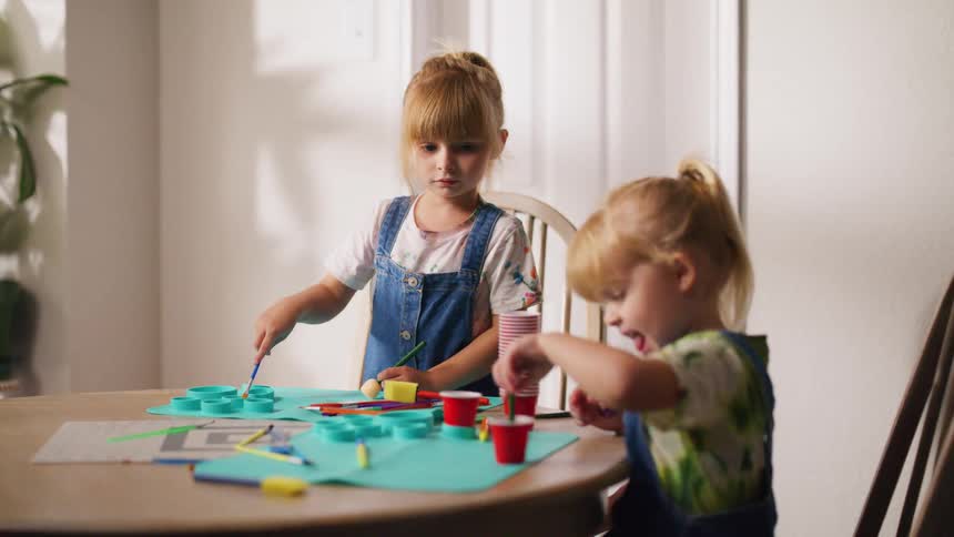 Two adorable young sisters concentrate on their arts and crafts project while painting together at the dining table