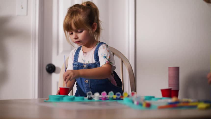 A cute young girl in overalls concentrates on painting during an arts and crafts session at a table in her home