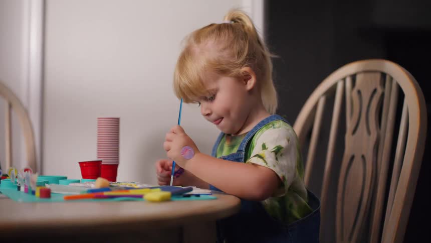 A cute toddler with her tongue out in concentration paints with a brush while sitting at a dining table