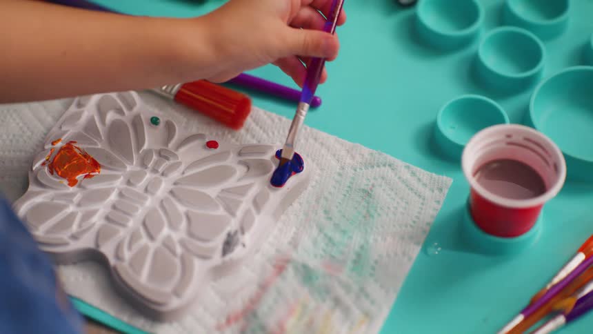 A child's hand carefully paints a butterfly-shaped plaster mold with a brush and blue paint during an arts and crafts session