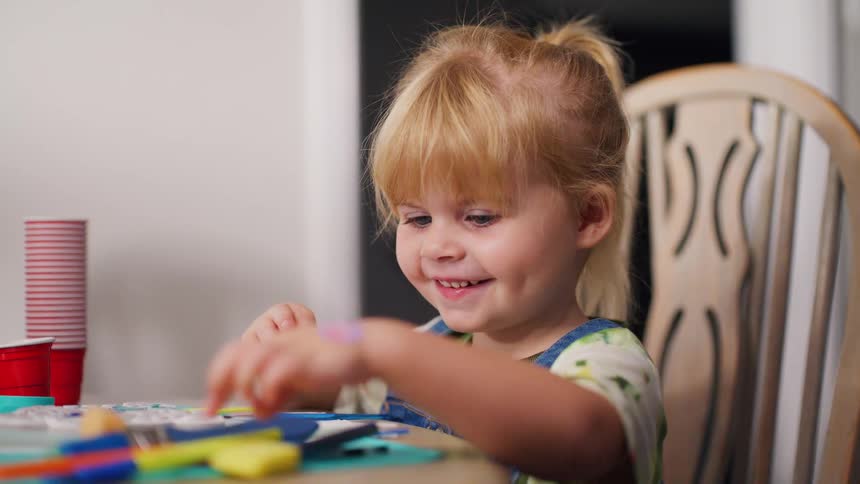 A happy toddler with blonde hair smiles while playing with paintbrushes at a table in slow motion