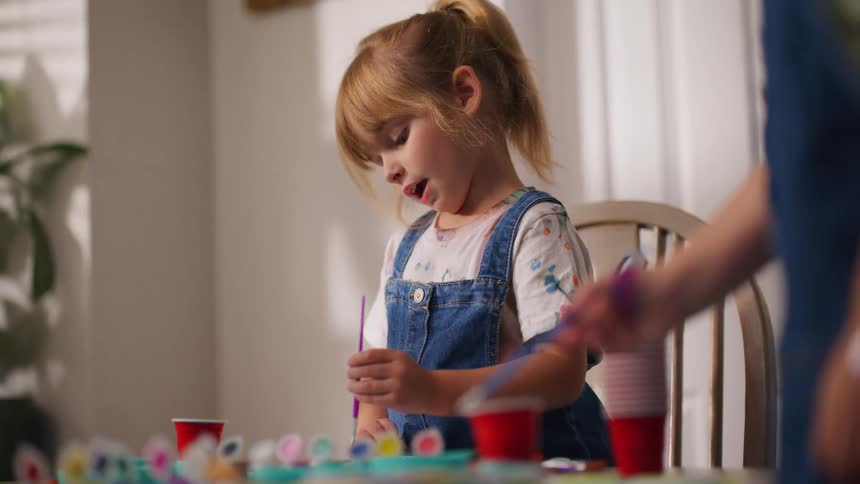 A focused young girl in overalls carefully paints at a table during an arts and crafts session at home