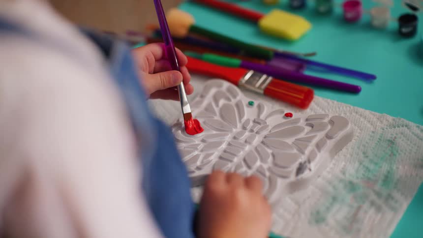 A close-up over-the-shoulder view of a child's hands carefully painting a plaster butterfly craft with red paint