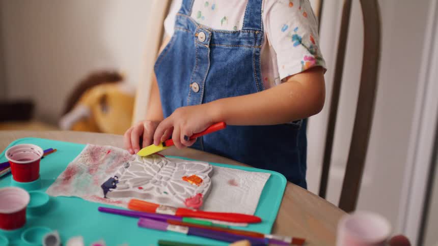 A creative young child in denim overalls sits at a table and carefully paints a butterfly craft