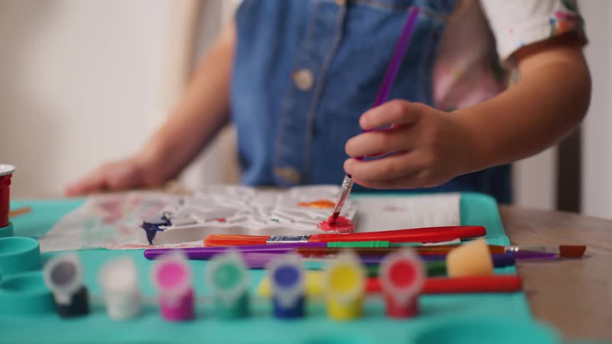 A close-up on a young child's hand as they use a small paintbrush to apply red paint to an art project, with colorful paints in the foreground