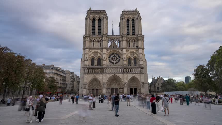 A long exposure timelapse shows crowds of blurred people moving through the plaza in front of the historic Notre Dame Cathedral in Paris, France