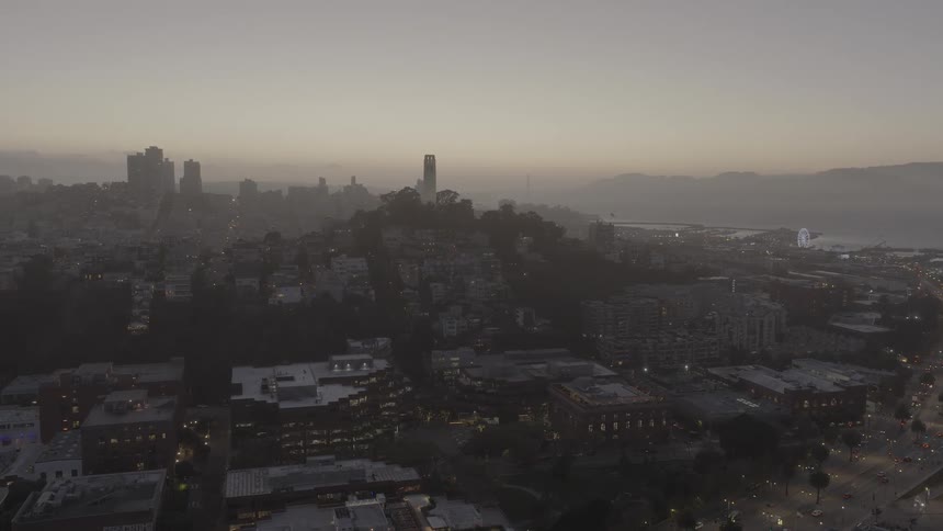 A slow aerial pan reveals the hazy San Francisco cityscape at dusk, with Coit Tower perched on a hill and the city lights illuminating the streets below