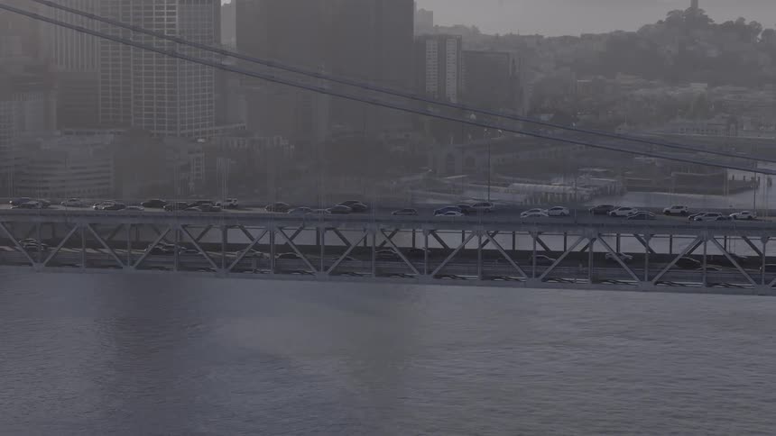 An aerial view of traffic flowing on the San Francisco-Oakland Bay Bridge with the city skyline visible through the haze in the background