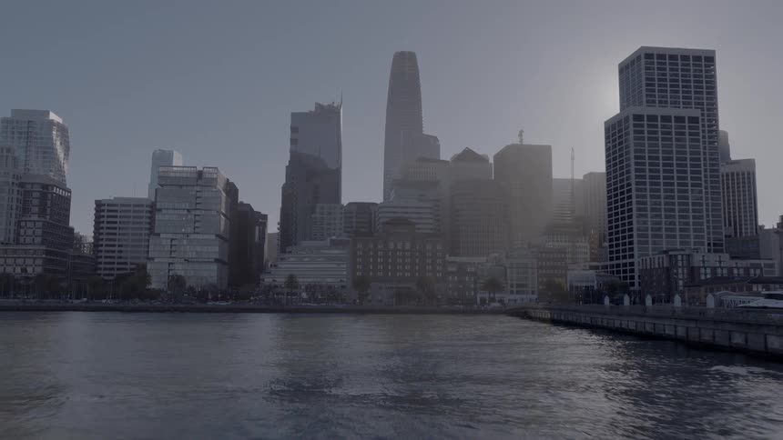 A slow aerial push-in shot of the hazy San Francisco skyline from over the water as the sun shines brightly