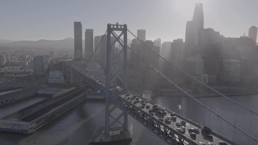 An epic aerial reveal of the San Francisco Bay Bridge, with traffic flowing into the hazy, sun-drenched city skyline