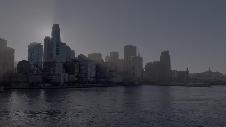 Aerial view flies under the San Francisco Bay Bridge at sunrise, revealing the city skyline with a beautiful sun flare over the water