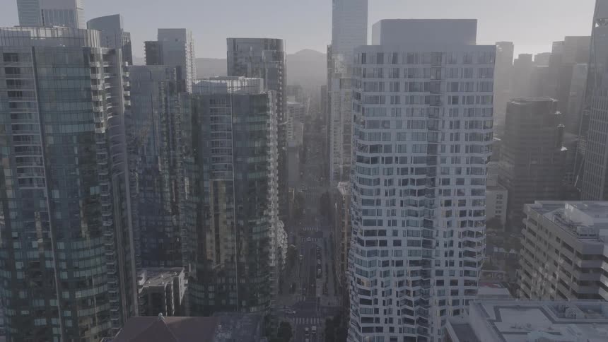 An aerial view reveals the urban canyon of a major city at sunrise, with the camera moving forward between tall skyscrapers and modern buildings