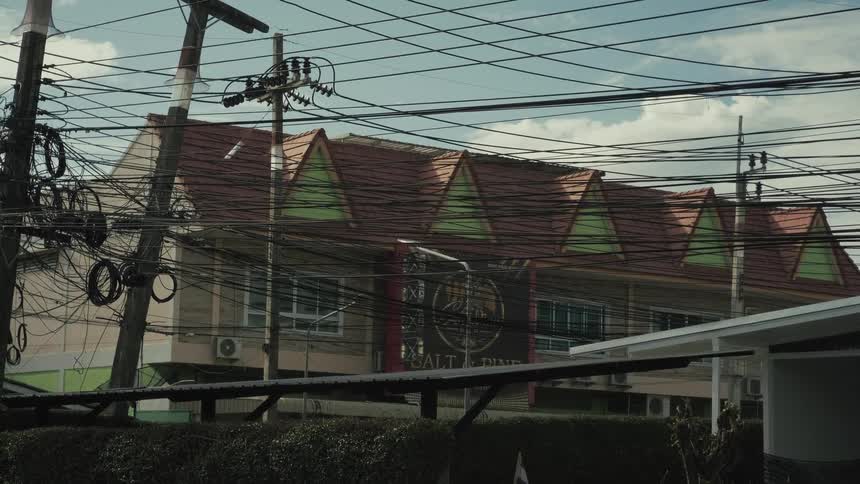 A slow zoom-out reveals a building with a red-tiled roof through a dense and chaotic web of electrical power lines in Thailand