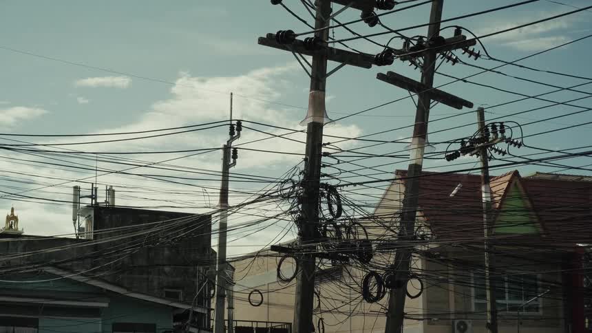 A slow tilt down on a utility pole reveals a chaotic mess of electrical wires and communication cables in a Thai city