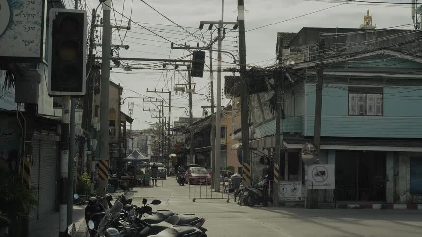 A static shot of a quiet intersection in a Thai town with parked motorbikes, traditional buildings, and a tangle of overhead wires