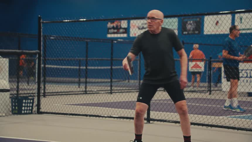 An elderly man focuses intently as he hits a forehand shot while playing pickleball on an indoor court