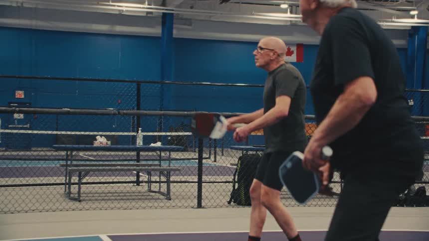Two senior men compete in a game of pickleball on an indoor court, captured in slow motion from a side angle