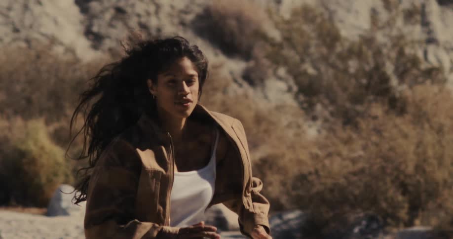 A determined woman with wind-blown hair runs in slow motion through a sunlit, arid desert landscape