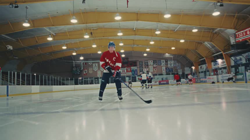 A male hockey player in a red jersey stickhandles a puck while skating towards the camera in slow motion in an indoor arena