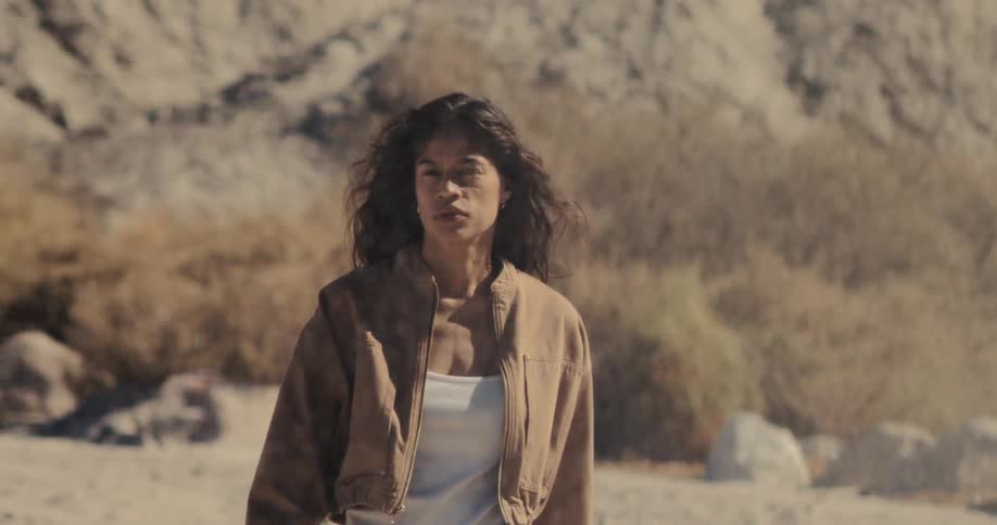 A confident woman stands with a serious expression as dust and sand blow around her in a desolate, arid landscape