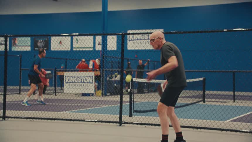 An elderly man serves the pickleball in slow motion while playing a match on an indoor court
