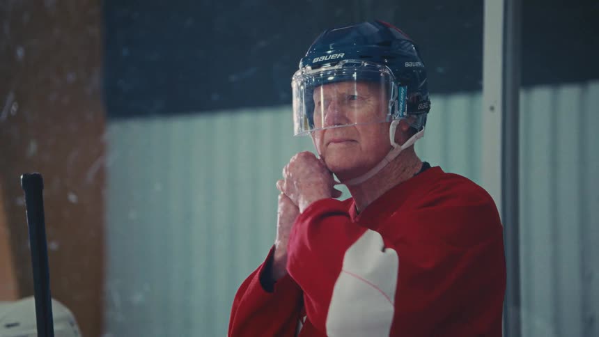 An elderly hockey player in a red jersey puts on his helmet before stepping onto the ice from the player's bench