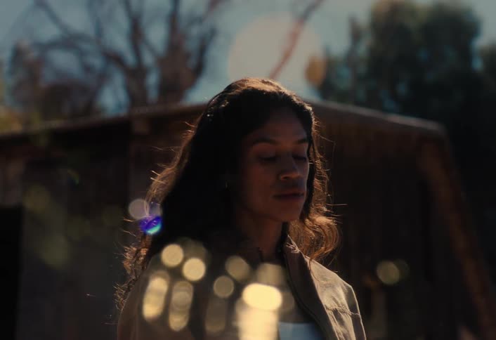 A serene woman with curly hair closes her eyes to feel the warm sun on her face in a peaceful rural setting