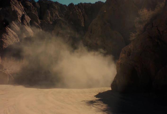 A thick cloud of dust billows through a desolate, rocky desert canyon on a sunny day
