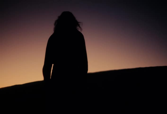 Slow motion silhouette of a person with windblown hair walking over a hill against a sunset sky