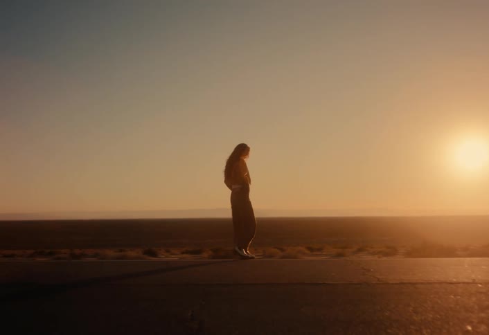 A woman walks and stops on a remote desert road, silhouetted by the warm glow of the rising sun