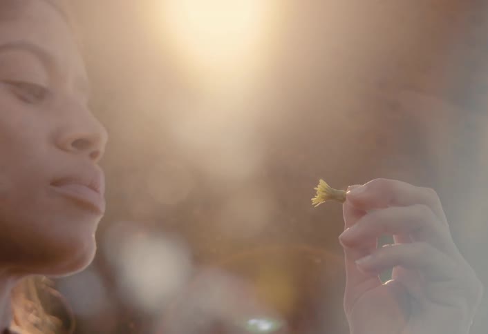 A serene woman finds a moment of peace while holding a small flower as sunlight flares and smoke drifts by