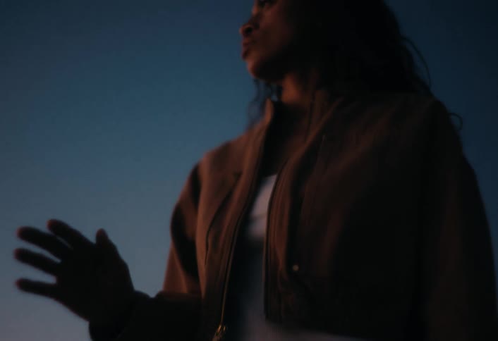 A pensive young woman in a jacket looks away and then down as she stands silhouetted against the evening sky
