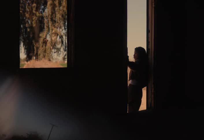 A woman stands silhouetted in a rustic doorway, looking out from a dark room into the sunny countryside